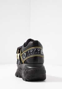 Svart sneaker med tjock sula, med gula accenter och vit märkessnodd. Strukturerat material, vadderad krage och framträdande mönster på sulan.