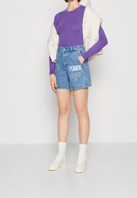 Persona che indossa un top a maniche lunghe viola a coste, pantaloni shorts in denim blu con la scritta "PEACE CHANCE", sneakers bianche e un gilet imbottito beige chiaro.