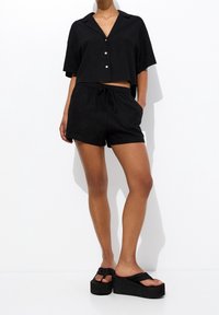 Camisa negra corta con botones y mangas cortas, combinada con pantalones cortos negros con cordón. Calzado compuesto por sandalias negras de plataforma gruesa.