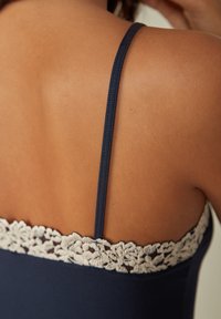 Vêtement bleu marine avec des fines bretelles, doté d'une garniture en dentelle crème en haut. Texture lisse sur le corps, montrant un gros plan du dos.