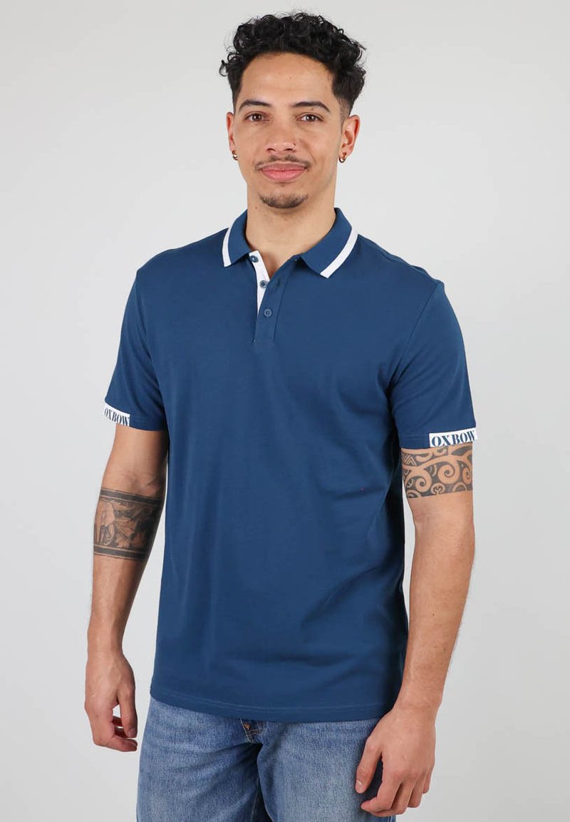 Polo shirt marine en coton, avec un col blanc et des finitions blanches aux manches, et un accent logo sur la manche gauche. Coupe classique.