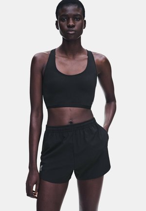 Frau trägt schwarzen Sport-BH und schwarze Sportshorts und steht mit einer Hand in der Tasche vor einfachem Hintergrund.