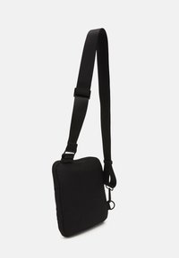Svart axelväska med fyrkantig form, tillverkad av texturerad tyg. Har en justerbar rem och ett dragkedjehölje på toppen.