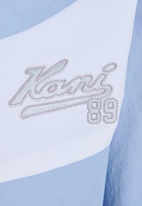 Lyseblå og hvit tekstil med brodert "Kani"-logo og "89" i sølvtråd. Glatt tekstur, sporty design, moderne fargeblokkstil.