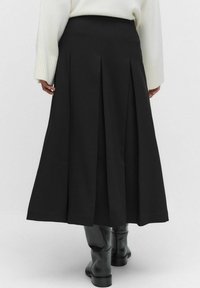 Sort plisseret midi-nederdel iført sorte knæhøje støvler og en hvid langærmet sweater, vist fra ryggen.