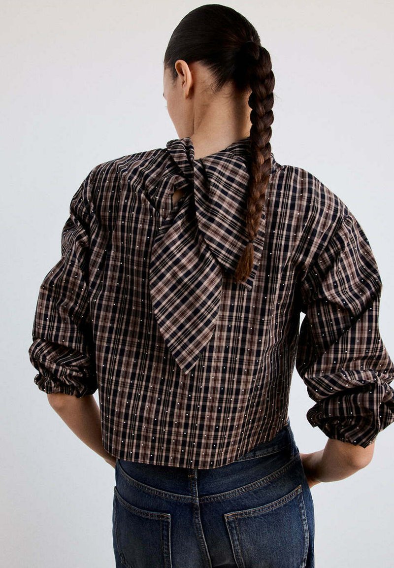 Femme avec une longue tresse portant un chemisier à carreaux marron et noir avec des manches bouffantes et un grand nœud dans le dos, associé à un jean foncé.