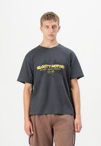 Un tânăr îmbrăcat cu un tricou gri închis pe care scrie în galben "Velocity Motors" și are steaguri în carouri încrucișate, asortat cu pantaloni maro.