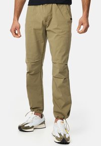 Khaki lightweight pants with a tapered design, elastic cuffs, and subtle stitching details. Worn with sporty white and green sneakers.