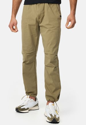 Khaki lightweight pants with a tapered design, elastic cuffs, and subtle stitching details. Worn with sporty white and green sneakers.