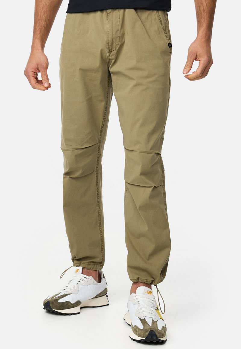 Khaki lightweight pants with a tapered design, elastic cuffs, and subtle stitching details. Worn with sporty white and green sneakers.
