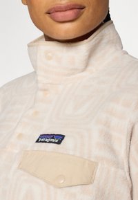 Beige Fleece-Pullover mit geometrischer Textur, hohem Kragen, vorderen Knöpfen und einer cremefarbenen Tasche mit einem blauen und roten Patagonia-Logo.