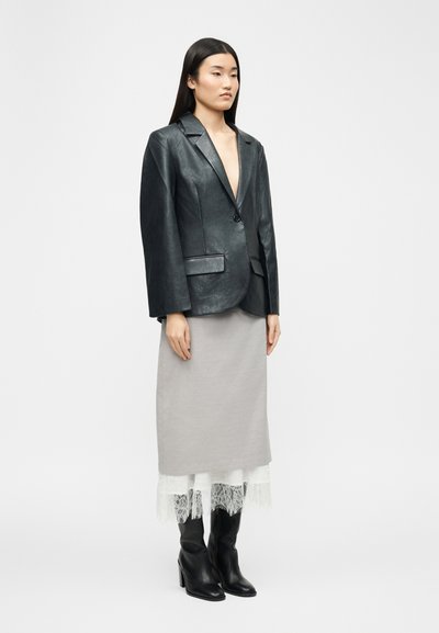 Γυναίκα που φοράει μαύρο δερμάτινο σακάκι, γκρι μίντι φούστα με λευκή δαντέλα και μαύρες μπότες με τακούνι, στέκεται μπροστά σε ένα απλό φόντο.