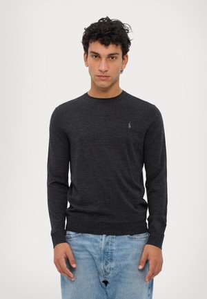 Temno siva dolgimi rokavi pulover iz mehkega blaga, s krojnim ovratnikom in majhnim vezenim logotipom na prsih. Kombiniran z svetlo modrimi kavbojkami.