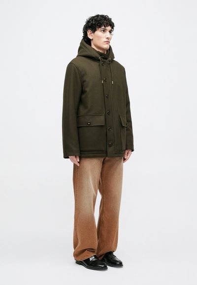 Oliivinvihreä hupullinen takki, jossa napit, kaksi etutaskua ja kiristysnaru. Yhdistetty beigen värisiin leveälahkeisiin housuihin ja mustiin kenkiin.