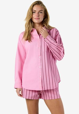 Ung kvinne iført en rosa skjorte med knapper, med én ensfarget og én stripete side, kombinert med matchende rosa stripete shorts, stående foran en hvit bakgrunn.