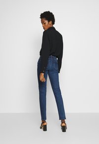 Camisa negra de manga larga, vaqueros de denim azules, de corte ajustado, de alta cintura, con bolsillos traseros y costuras claras, combinados con sandalias negras de tacón.