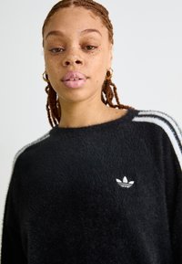 Sweatshirt preto felpudo com riscas horizontais brancas nas mangas e pequeno logótipo branco no peito. Decote redondo e textura suave.