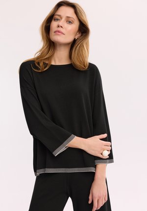 Kobieta o jasnobrązowych włosach, ubrana w czarną bluzkę z długim rękawem z białym paskowanym wykończeniem i pasujące czarne spodnie, pozująca z założonymi rękami.