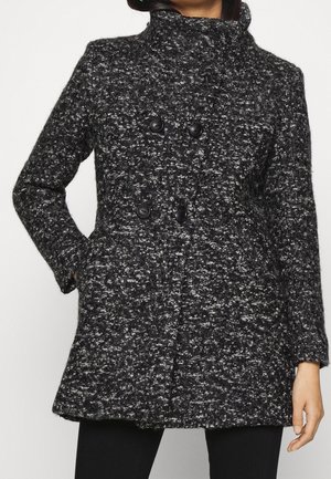 Femme portant un manteau en laine mouchetée noir et blanc avec poches avant et fermeture double à boutons noirs, debout avec une main dans la poche.