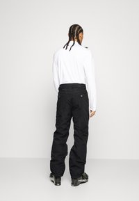 Zwarte sneeuwbroeken met een ruime pasvorm en elastische boorden, gecombineerd met een lange mouwen witte shirt. Schoenen hebben zwarte en neon details.