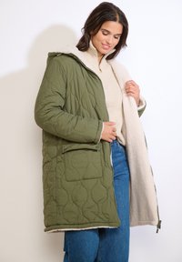 Žena v prešívanom olivovo zelenej bunde s fleecovou podšívkou, béžovom roláku a modrých džínsach, ktorá jednou rukou drží kabát otvorený.