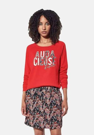 Sweat-shirt rouge à manches longues avec "AUDACIEUSE" en lettres à motifs, associé à une jupe florale noire avec des nuances d'orange et de bleu.
