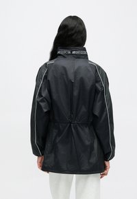 Μαύρο αδιάβροχο μπουφάν με ψηλό γιακά, λευκές ραφές και ρυθμιζόμενο κορδόνι στη μέση. Διαθέτει το λογότυπο "NIKE SHOX" στον γιακά.