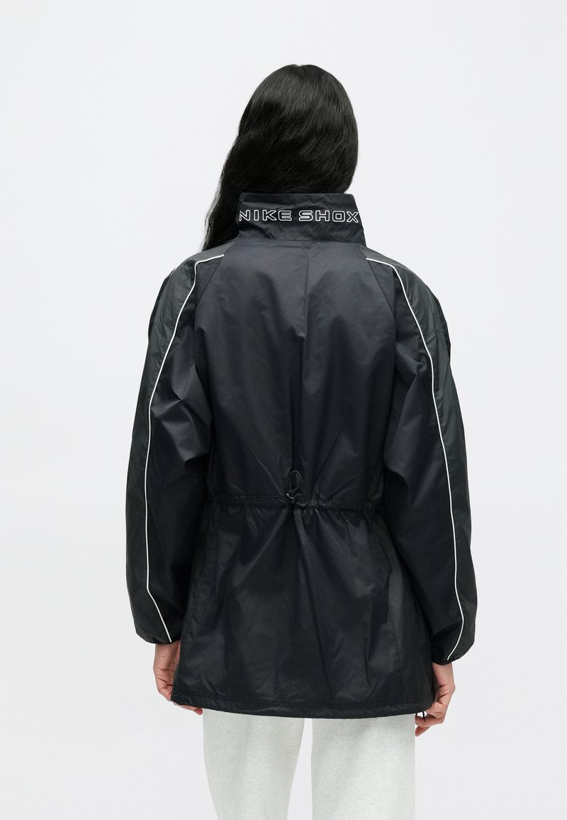 Schwarze Windbreaker-Jacke mit hohem Kragen, weißen Paspelierungen und verstellbarem Kordelzug in der Taille. Mit "NIKE SHOX"-Logo am Kragen.
