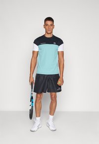 Hellblaues Sportshirt mit marineblauen und weißen Akzenten, kombiniert mit schwarzen Shorts. Hält einen Tennisschläger und einen gelben Ball.