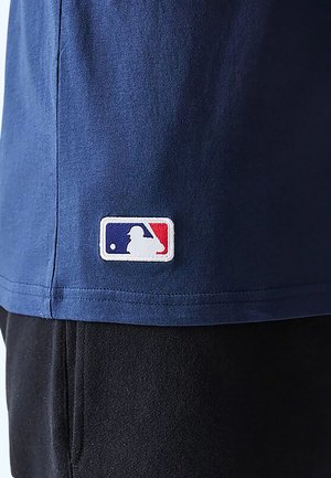 Blue shirt hem with a stitched MLB logo patch showing a white baseball batter silhouette on red and blue background above black pants.
