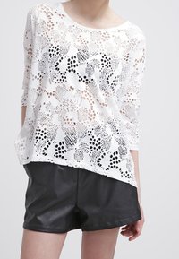 Weißes Spitzentop mit floralem Lochmuster, Dreiviertelärmel, lockere Passform, getragen über schwarzen Leder-Shorts, mit rundem Halsausschnitt.