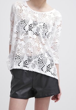 Top bianco in pizzo con motivo a fiori, maniche a tre quarti, vestibilità rilassata, indossato sopra pantaloni di pelle neri, con scollatura rotonda.