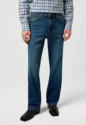 Jean en denim bleu foncé avec une coupe droite, présentant une fermeture à glissière et bouton, cinq poches et des accents de couture subtils.