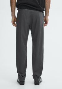 Pantalons gris avec une coupe ajustée, dotés d'une poche arrière unique et d'une texture de tissu lisse, associés à des chaussures noires.
