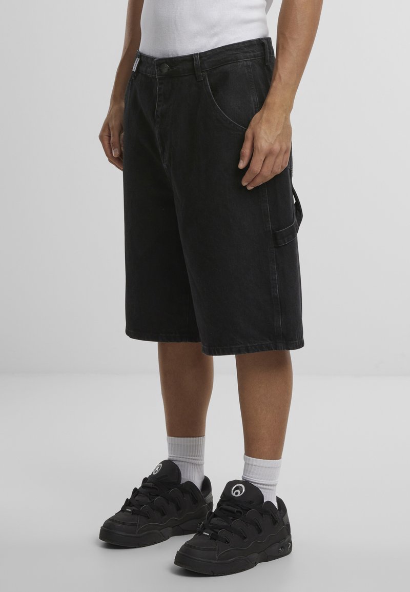 Pantalones cortos de denim negros con un ajuste suelto, con bolsillos frontales y un bolsillo lateral tipo cargo. Llevados con zapatillas negras y calcetines blancos.