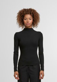 Čierna košeľa s dlhými rukávmi a vysokým golierom, vyrobená z hladkej látky. Tesne prilieha k telu s elegantným tvarom, nosená s tmavými nohavicami.