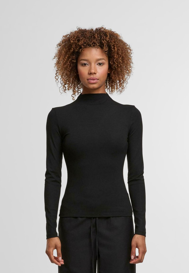 Čierna košeľa s dlhými rukávmi a vysokým golierom, vyrobená z hladkej látky. Tesne prilieha k telu s elegantným tvarom, nosená s tmavými nohavicami.