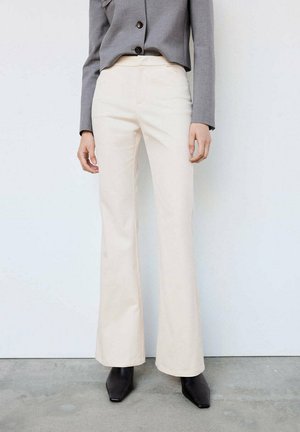 Persona que lleva pantalones acampanados crema de cintura alta, chaqueta corta gris y botas negras de punta afilada, de pie sobre un suelo de hormigón.