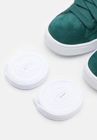 Gröna mockaskor med vit gummisula, med rund vita snören prydligt hoprullade bredvid dem på en neutral bakgrund.