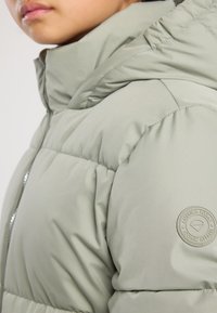 Plano cercano de una persona que lleva una chaqueta acolchada gris claro con un cuello alto y un logo circular en la manga.