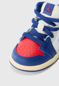 Zapatillas de cuero azules y rojas con una superposición de puntera perforada, parte superior de textil blanca, cordones azules y etiqueta con el logo en la lengüeta.