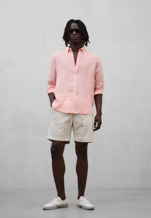 Mann mit Dreadlocks, der schwarze Sonnenbrille, ein hellrosa Leinenhemd, beige Shorts und weiße Turnschuhe trägt und vor einer grauen Wand steht.