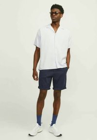 Chemise blanche à manches courtes, short à motif bleu marine, baskets blanches et chaussettes bleues. Des lunettes de soleil complètent ce look décontracté.
