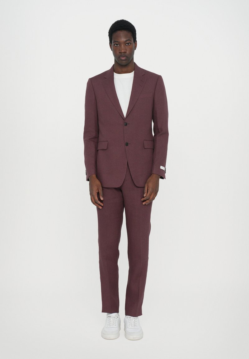 Bordeaux farbener Anzug mit tailliertem Blazer und Hose, kombiniert mit einem weißen T-Shirt und weißen Sneakers. Glatte Textur, klassisches Design.