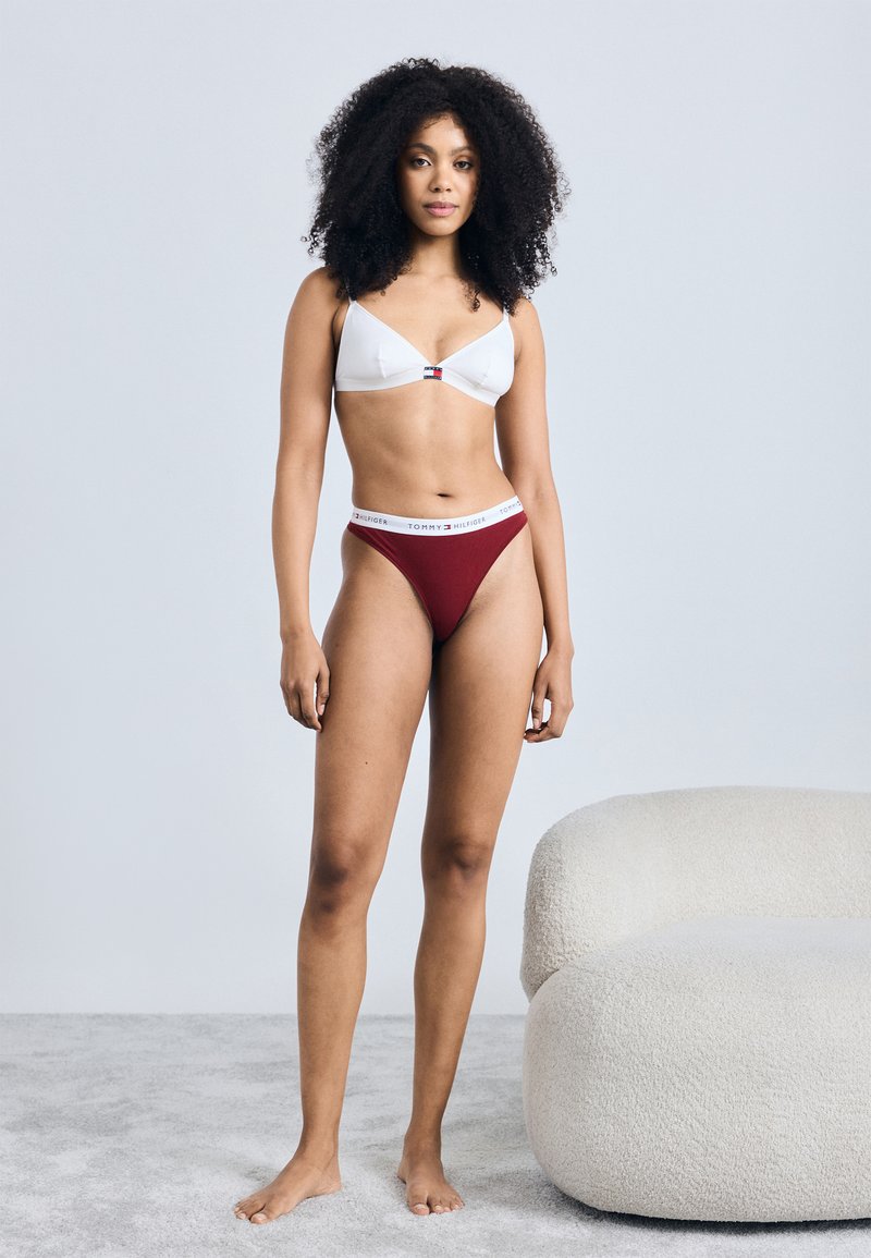 Balta trijstūra krūšturis ar nelielu logo detaļu, apvienots ar tumši sarkano tanga apakšveļu ar baltu zīmola jostu; modelis stāv basām kājām uz gaiši pelēka paklāja.