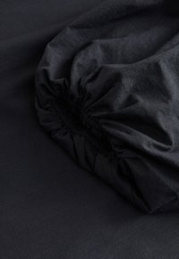 Coin de drap housse gris foncé avec bord élastique, montrant la texture du tissu et des coutures froncées sur une surface plane.