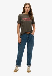 Camiseta de algodón gris con "Superdry Soho, Londres" en texto rosa, combinada con jeans de denim azul con los puños doblados y zapatillas beige.