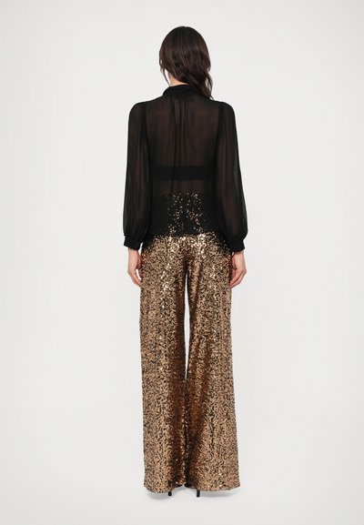 Bluză neagră transparentă cu mâneci lungi bufante, asortată cu pantaloni largi, cu talie înaltă, din sequinuri în nuanță metalică de bronz.