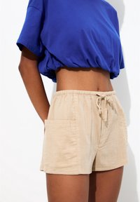 Shorts en coton beige avec une taille élastique et un cordon ajustable, dotés d'une poche latérale et d'une finition douce et texturée.