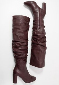 Botas de cuero burdeos por encima de la rodilla con un diseño arrugado, tacones altos anchos y cremallera lateral. Textura suave con detalles de costura.